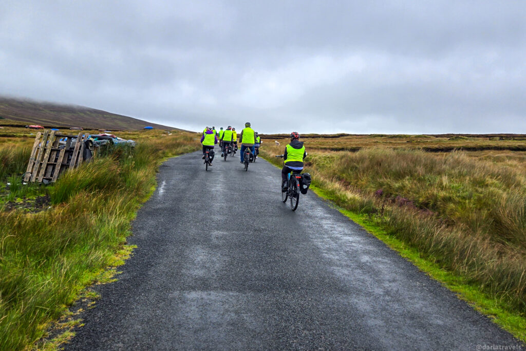 A group of cyclists wearing bright yellow vests rides away down a wet, narrow paved road through a grassy, moorland landscape under an overcast sky, in Co. Donegal, Ireland.