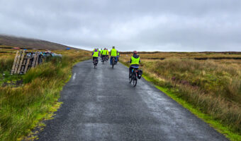 A group of cyclists wearing bright yellow vests rides away down a wet, narrow paved road through a grassy, moorland landscape under an overcast sky, in Co. Donegal, Ireland.