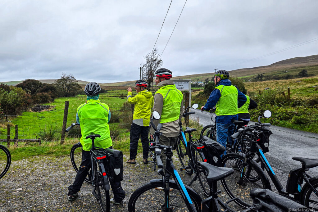 Group of cyclists and their e-bikes wearing bright safety vests pause by the roadside in a lush, green, hilly landscape under an overcast sky; in County Donegal, Ireland