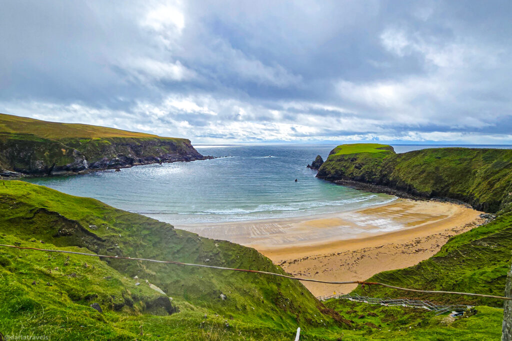 Silver Strand Beach, west coast of County Donegal, Ireland; secluded sandy cove framed by steep, lush green cliffs meeting a dramatic, overcast sea.
