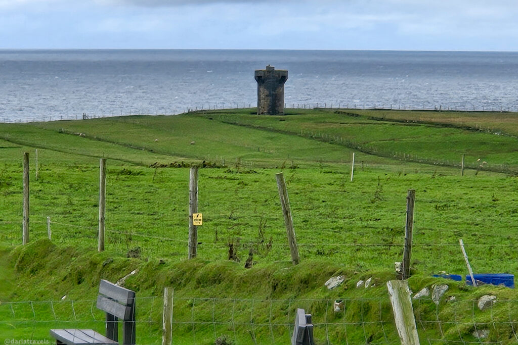 Malin Beg Signal Tower on Co. Donegal's west coast, a dark stone watchtower stands on a bright green, fenced coastal hill overlooking the grey, choppy ocean under an overcast sky.