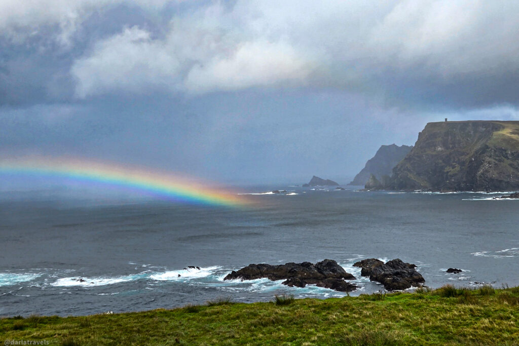 A vibrant rainbow arches over dark, churning ocean water near rugged sea cliffs under a stormy, gray sky, viewed from a grassy foreground; viewpoint in County Donegal, Ireland