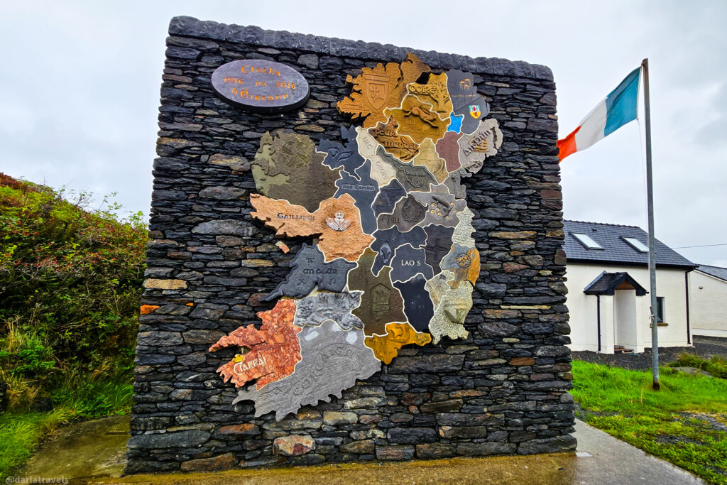 A multicolored stone relief map of the counties of Ireland mounted on a dark stacked stone wall, with an Irish flag waving nearby.
