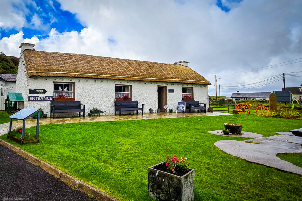 A white, traditional Irish cottage with a thatched roof and an "ENTRANCE" sign stands before a bright green lawn under a cloudy sky.