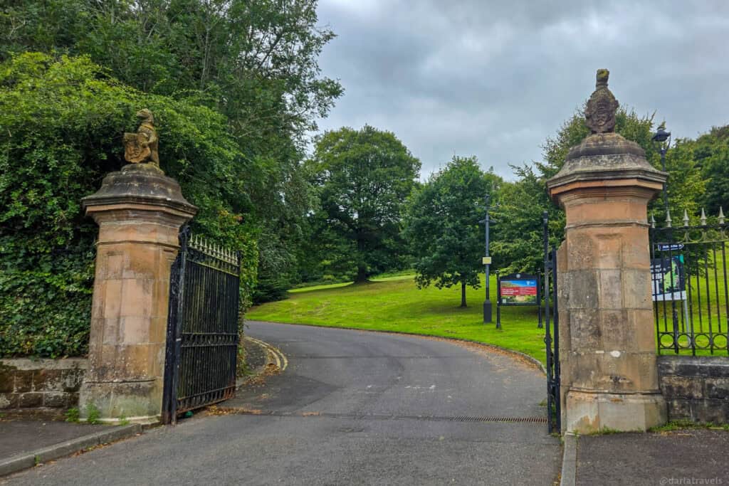 Stone entrance pillars and open iron gates at the lower entrance to Belfast Castle Estate, with a winding driveway leading uphill through green lawns and mature trees under a cloudy sky.