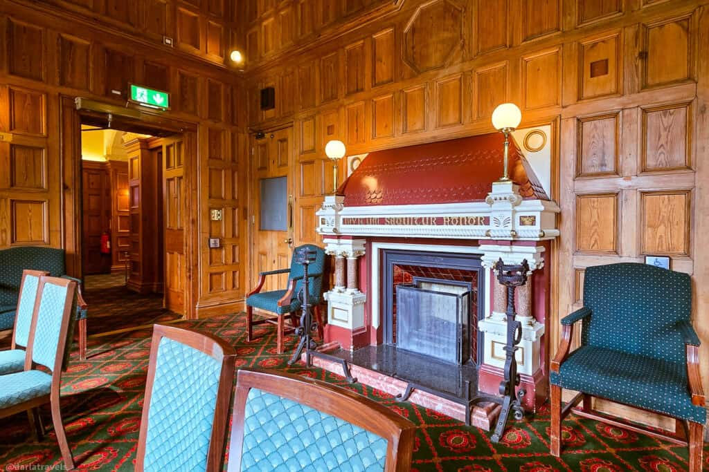 Wood‑paneled room inside Belfast Castle with an ornate red and cream fireplace, vintage green upholstered chairs, and patterned carpet set up for a small event.