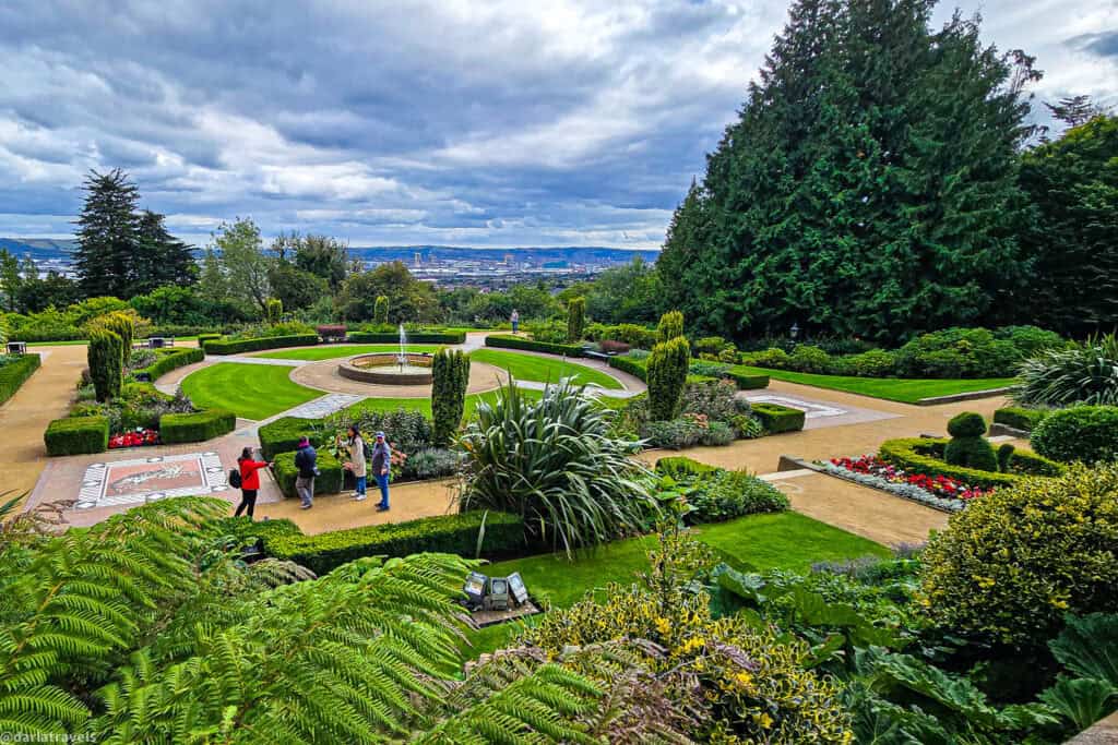 Panoramic view over Belfast from the formal terraces of Belfast Castle, with circular lawns, a central fountain, flower beds, and visitors walking through the Cat Garden.