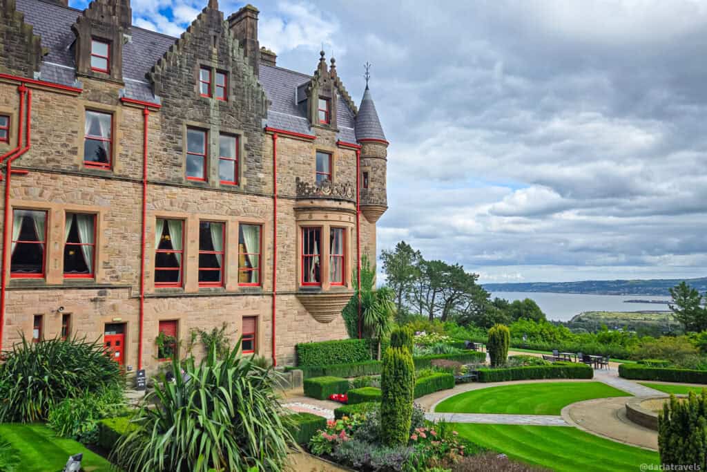Side view of Belfast Castle with its gabled roofline, red-trimmed windows, and formal gardens cascading down toward Belfast Lough in the distance.”