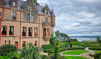 Side view of Belfast Castle with its gabled roofline, red-trimmed windows, and formal gardens cascading down toward Belfast Lough in the distance.”