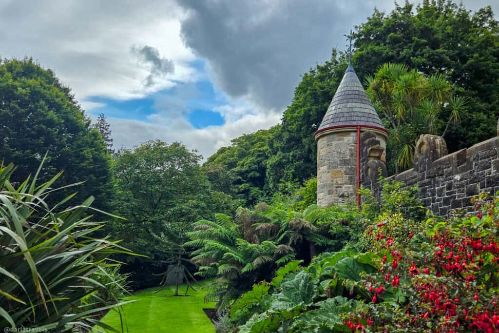 Stone turret and garden wall of Belfast Castle rising above lush green lawns and ferns, with dense trees and dramatic clouds framing a patch of blue sky.