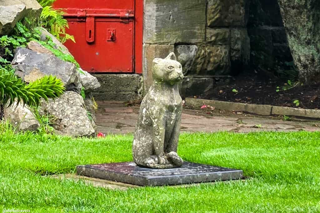 Memorial stone cat statue on a plinth engraved ‘In memory of Audrey Beggs, cat lover,’ with a cheerful cat tile and colorful flowers in the rock wall behind.