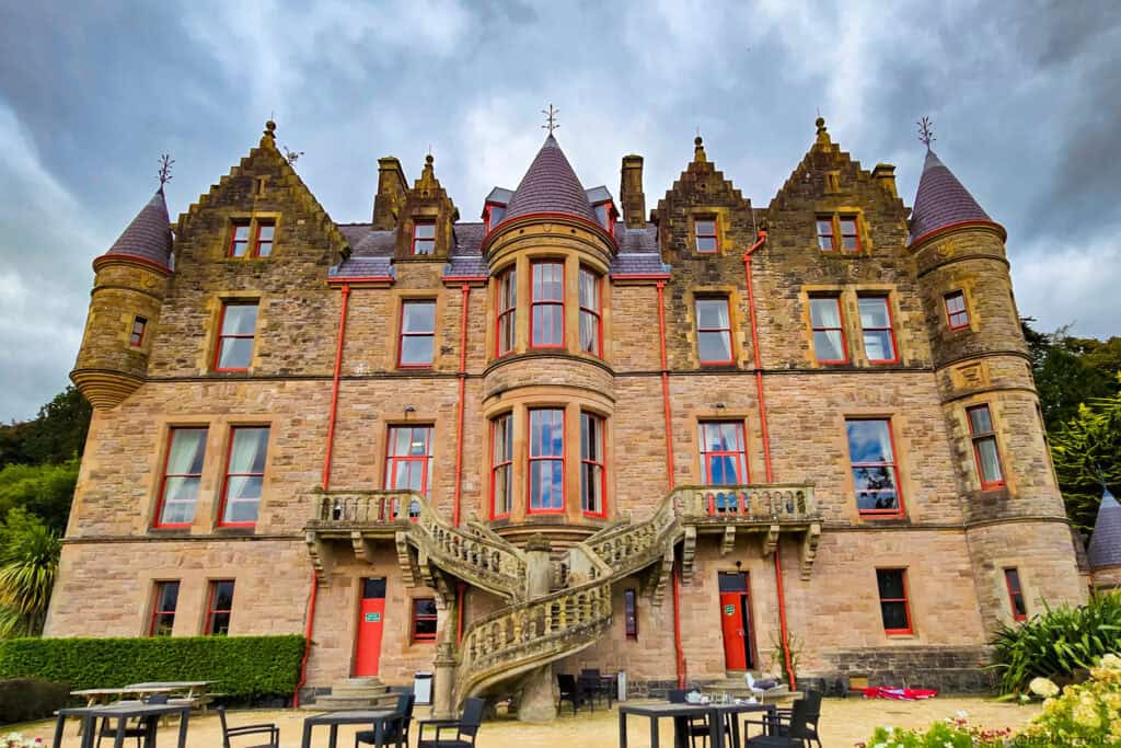 Rear facade of Belfast Castle in Scottish baronial style, with turrets, red-trimmed windows, and a dramatic double stone staircase above a terrace with outdoor tables.