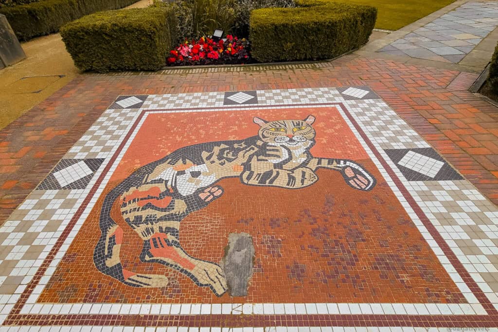 Colorful mosaic of a reclining tabby cat set into the paving of Belfast Castle’s Cat Garden, bordered by brick paths, low hedges, and bright red flowers.