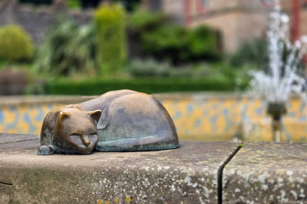 Close-up of a small bronze cat sculpture curled up and sleeping on the fountain ledge in Belfast Castle’s Cat Garden, with the tiled basin and water jets softly blurred behind