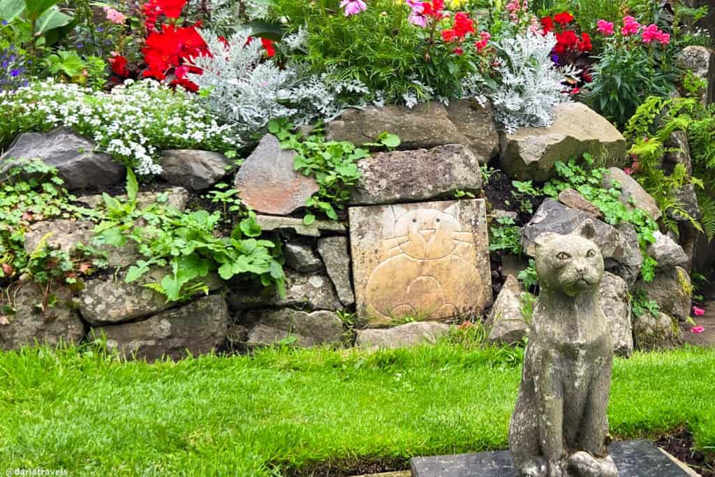 Rock garden in Belfast Castle’s Cat Garden with bright red and white flowers, a whimsical carved cat tile set into the stones, and a seated stone cat memorial on a small plinth
