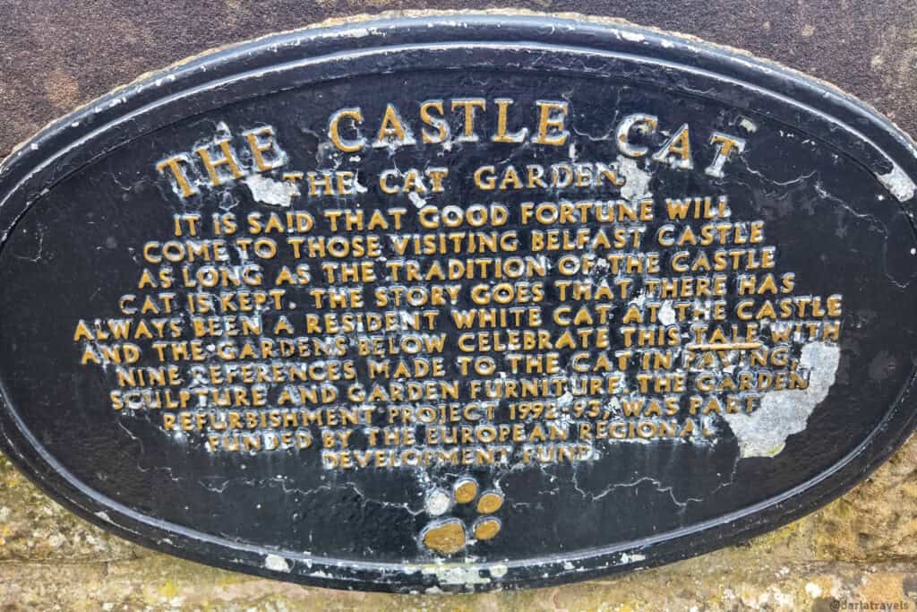 A close-up of the weathered black plaque in Belfast Castle’s Cat Garden explaining the legend of the resident white cat and the nine cat references, with a small golden paw print at the bottom.