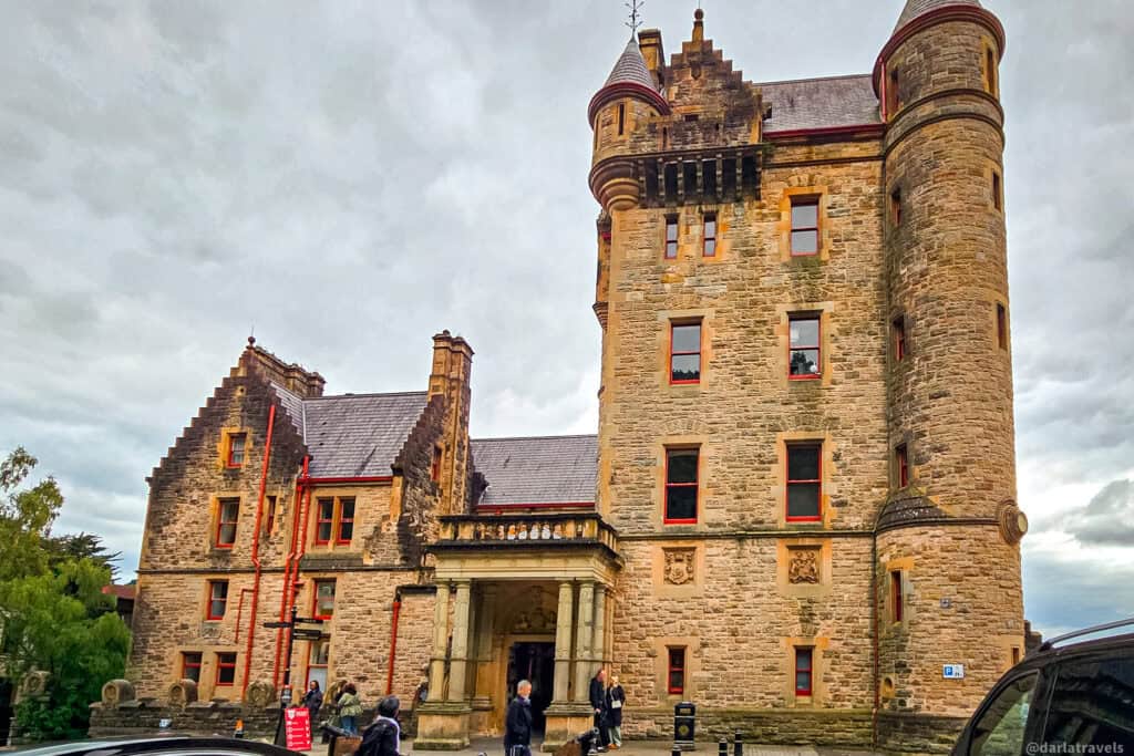 Front facade of Belfast Castle’s main entrance, showing the tall round tower, stone portico, and visitors arriving under a cloudy sky.