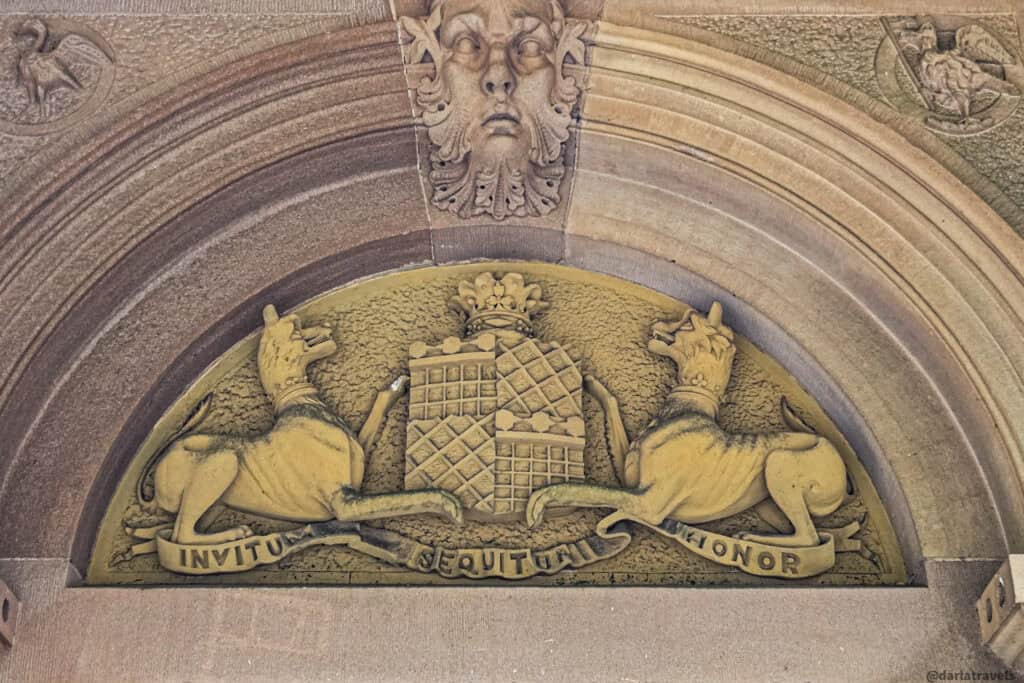 Stone coat of arms above the entrance to Belfast Castle showing two wolves supporting a shield and crown, framed by carved faces and decorative arches.