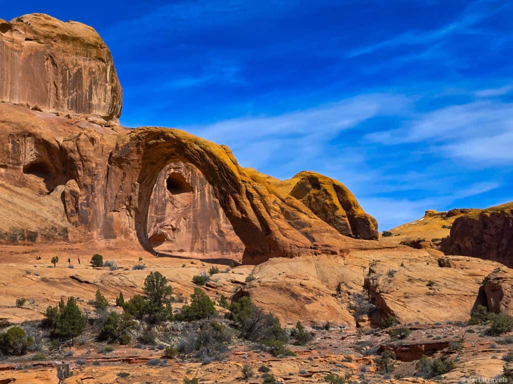 Wide view of Corona Arch and surrounding red rock cliffs under a bright blue sky near Moab, with tiny hikers visible on the desert floor.