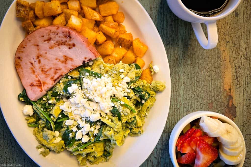Green eggs and ham breakfast plate with pesto scrambled eggs, feta, roasted potatoes, ham slice, fruit cup and black coffee on a rustic table