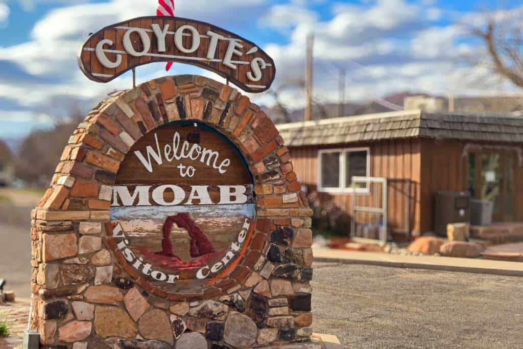 Stone “Coyotes Welcome to Moab Visitor Center” sign made of desert rock in front of a rustic brown building under a blue sky
