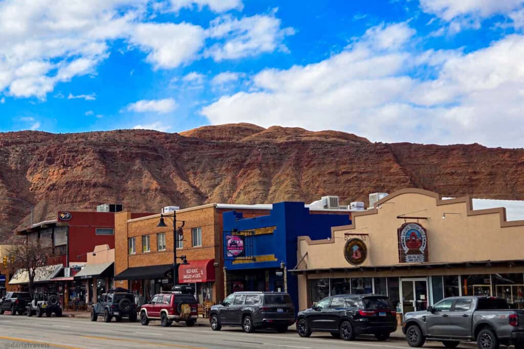 Downtown Moab Main Street with parked cars, colorful storefronts and red rock cliffs rising directly behind the buildings under a bright blue sky