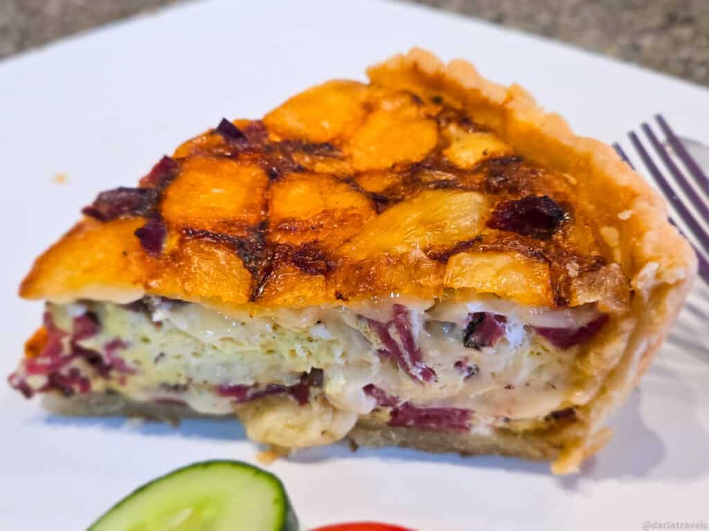 Close-up slice of pastrami and Swiss quiche on a white plate with cucumber garnish