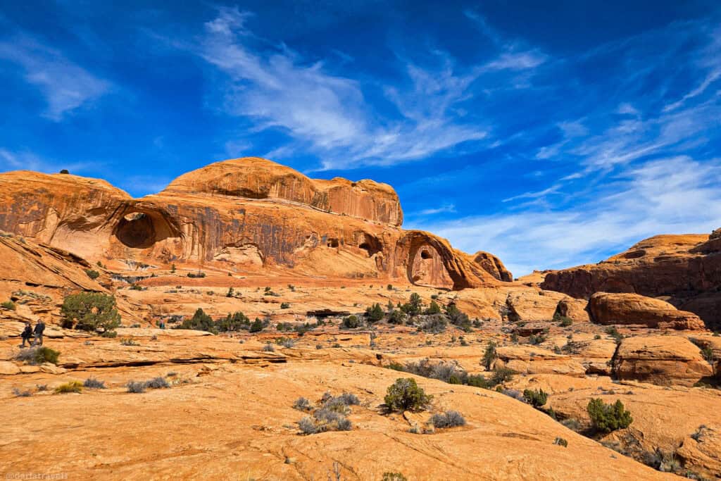 Hikers crossing slickrock below a broad sandstone arch. Corona Arch, Bowtie Arch.