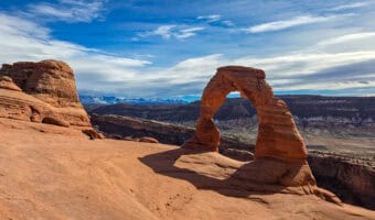 Delicate Arch rising above slickrock with snowy La Sal peaks on a short hike in Arches National Park.