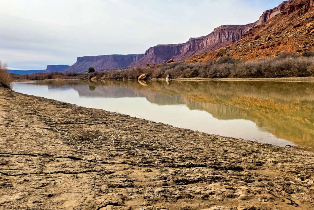 Calm river reflecting red cliffs beside a cracked desert shoreline along a quiet Moab‑area walk. Hittle Bottom