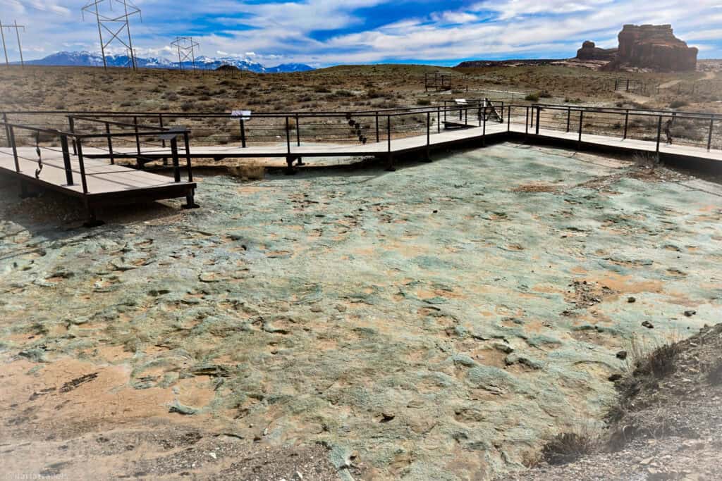Wooden walkway overlooking a broad field of dinosaur tracks with red cliffs and snowy peaks in the distance.