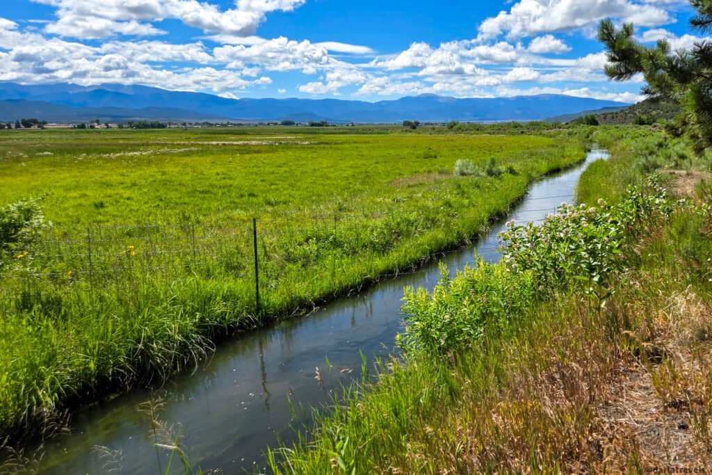 Green wetland meadow with flowing water and distant mountain range under a blue cloudy sky in San Luis Colorado