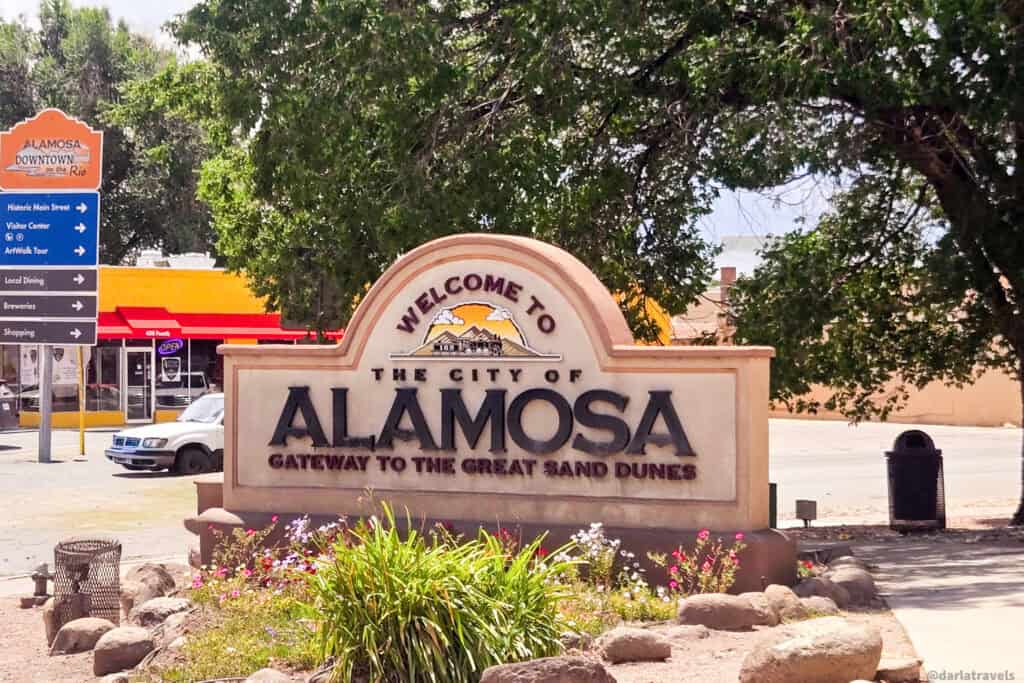 Welcome to Alamosa Colorado sign with Gateway to the Great Sand Dunes slogan on downtown street with tourist direction signs
