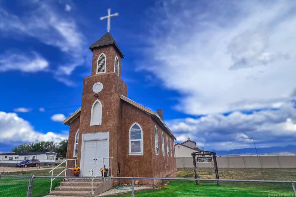 Brown adobe mission church with white bell tower and sign reading St. James Catholic Church in Blanca