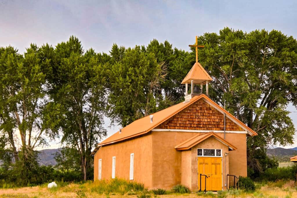 Rustic adobe mission church with yellow doors and copper roof under cottonwood trees in San Luis Valley
