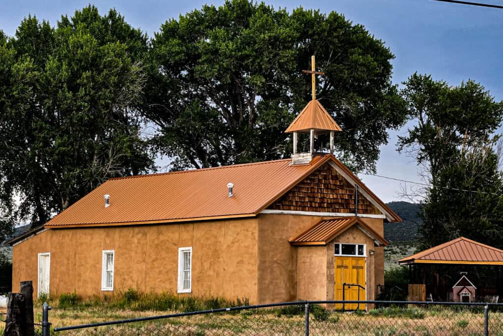 Tan adobe mission church with copper roof, yellow doors, and bell tower with cross in Costilla County