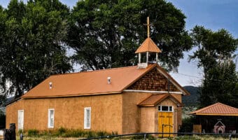 Tan adobe mission church with copper roof, yellow doors, and bell tower with cross in Costilla County