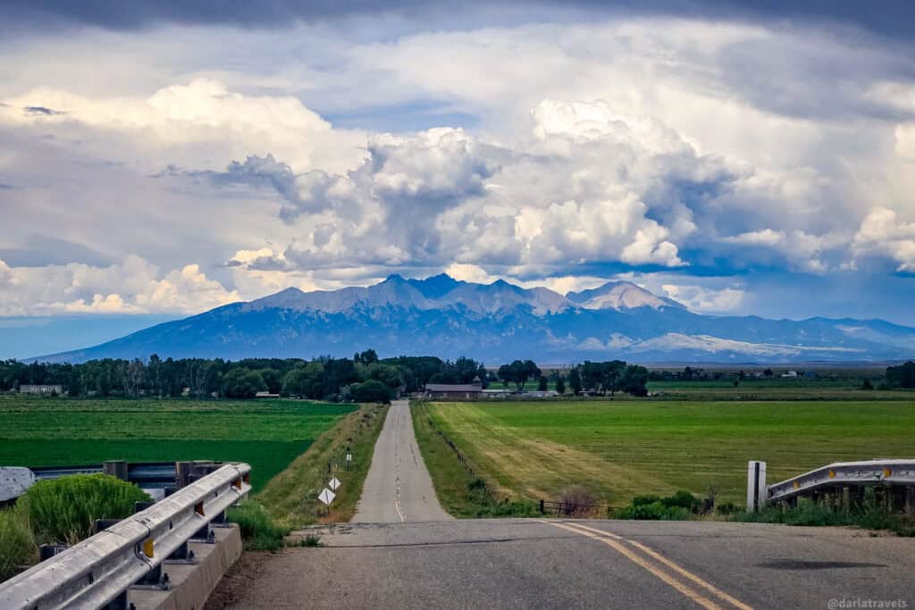 Scenic Colorado country road with green fields and majestic mountain range under heavy cloud cover in San Luis Valley