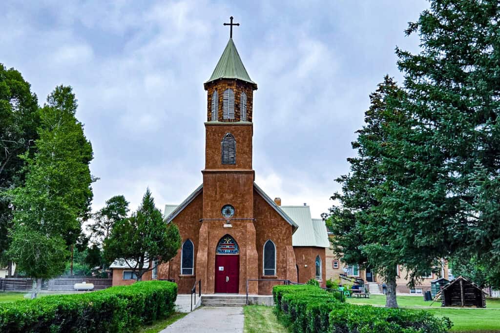 Adobe parish church with green bell tower and rose window surrounded by evergreen trees in Colorado