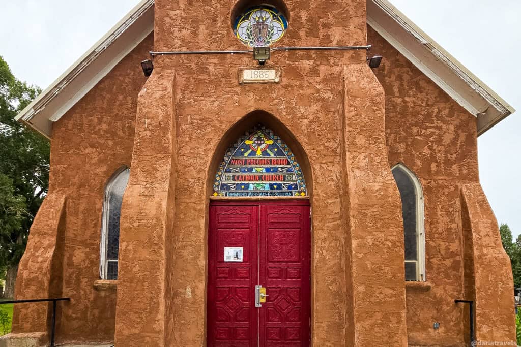 Adobe Catholic church with red doors, 1886 date, and Most Precious Blood stained-glass transom in San Luis Valley