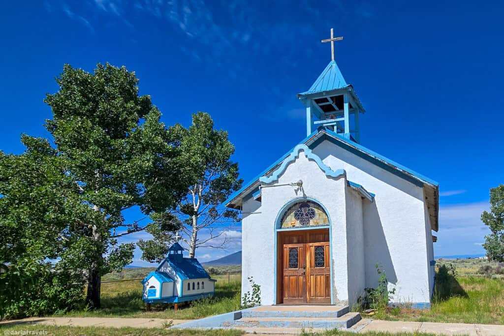 White adobe mission church with blue trim, blue bell tower, and miniature processional church in Garcia, Colorado