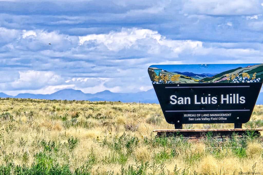 San Luis Hills Bureau of Land Management sign in golden grassland with distant mountains under cloudy sky