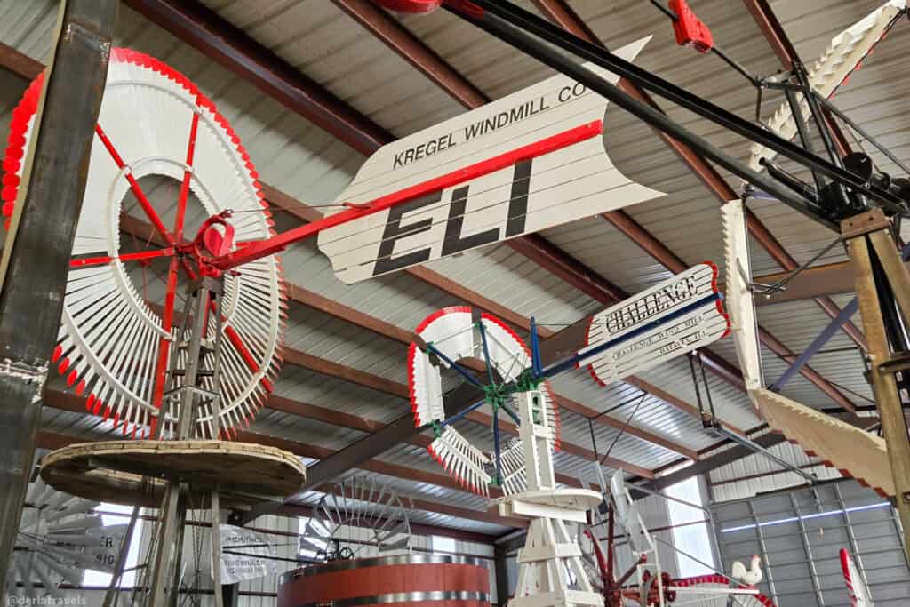 Colorful restored windmills and tail vanes fill an indoor barn museum, including one labeled “ELI”