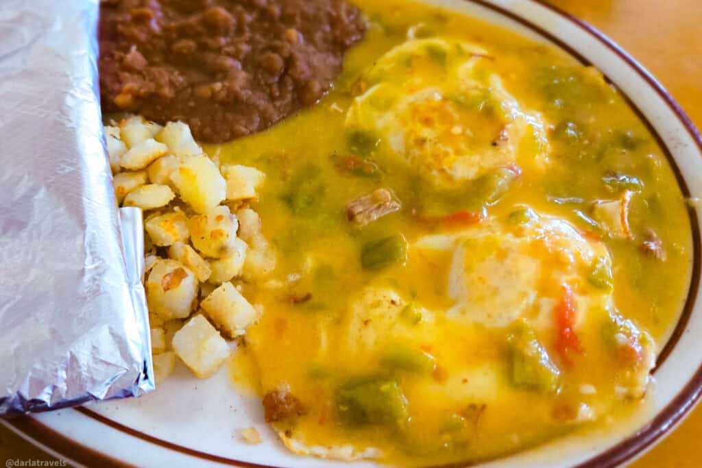 Breakfast plate with eggs smothered in green chile, diced potatoes, refried beans, and a foil-wrapped tortilla