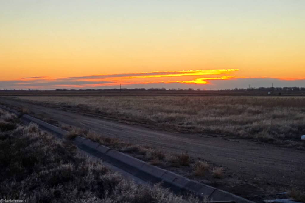 Soft orange sunrise glows over flat prairie fields and a dirt road on the high plains