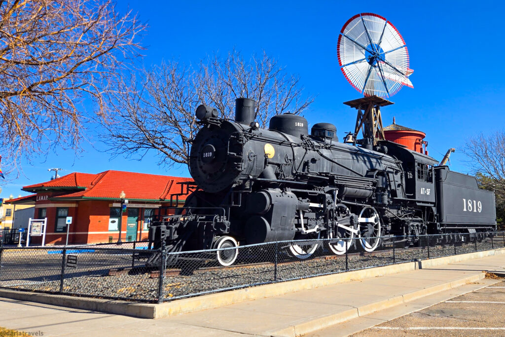 Historic black steam locomotive 1819 displayed outside Lamar Welcome Center with a vintage windmill behind it
