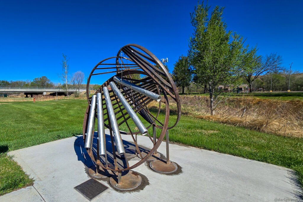 Metal sculpture with cylindrical tubes sits on a concrete pad with a grassy field and bridge in the background under a bright blue sky.