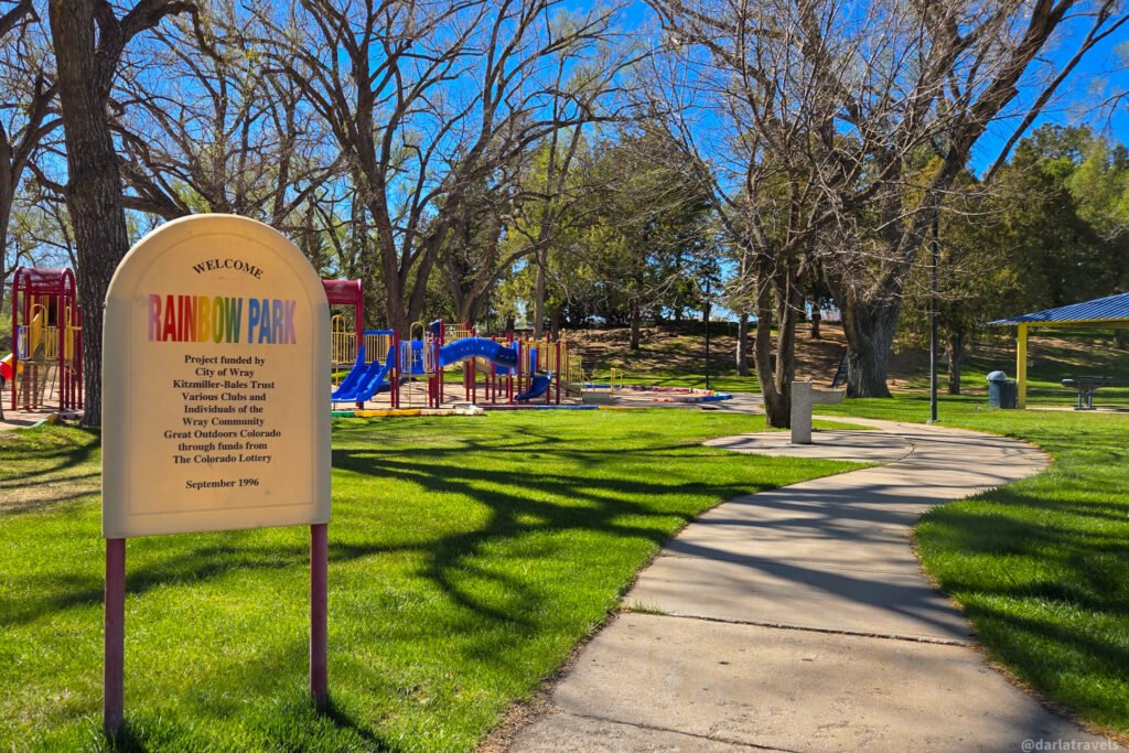 Welcome sign at Rainbow Park next to a playground with blue slides and a winding sidewalk under trees.