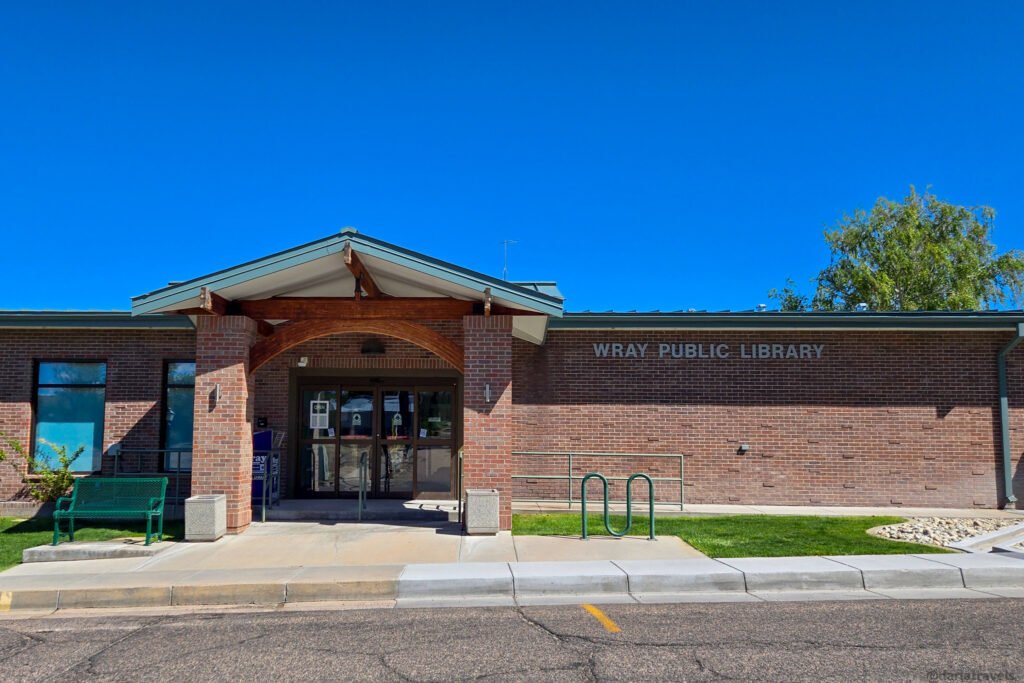 Wray Public Library entrance with brick columns, glass doors, and a green bench.