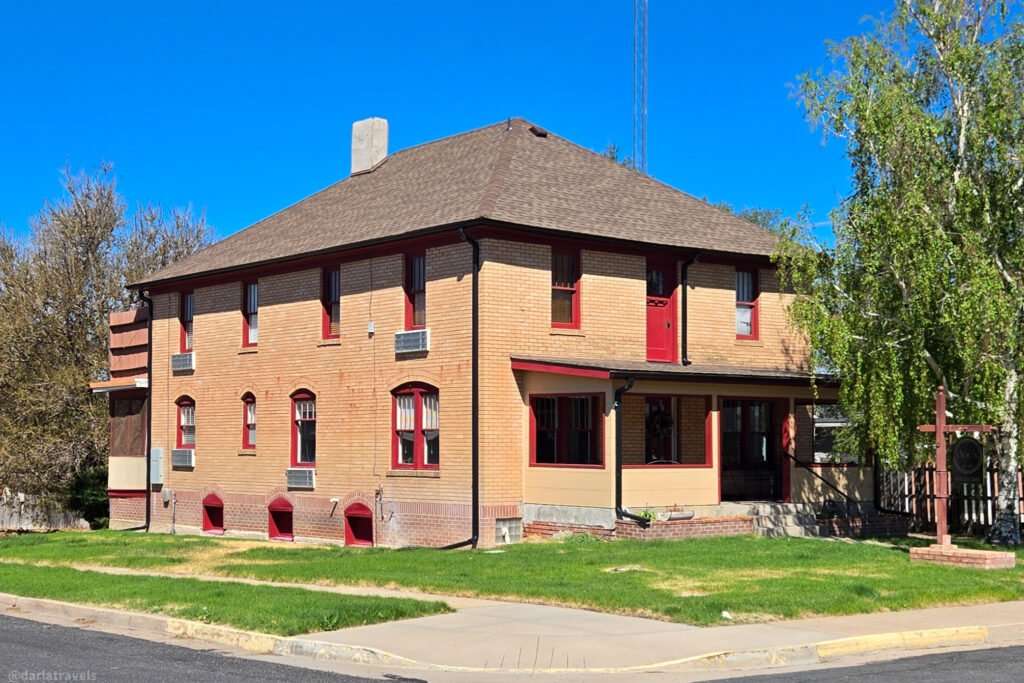 A two-story brick building with red trim, windows, and doors stands on a corner lot with green grass and a sidewalk.